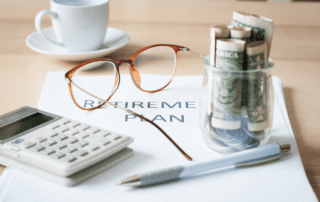 A desk with a coffee cup on it with a packet of paper infront of it that has the words "retirement plan" on it. The packet of paper has eye glasses, a pen, a calculator, and a jar with dollar bills rolled up in it on top of it.